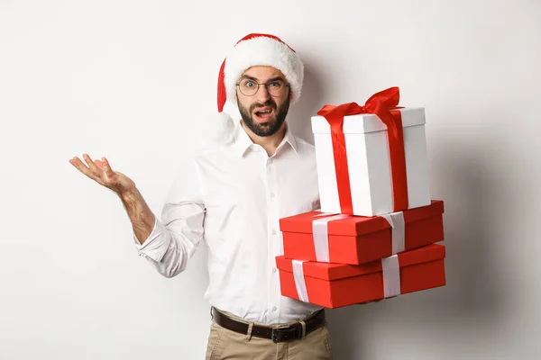 Wesołych Świąt, świąt. Człowiek patrząc zdezorientowany, trzymając prezenty Boże Narodzenie, wzrusza zagadką, stojąc w kapeluszu Mikołaja na białym tle — Zdjęcie stockowe