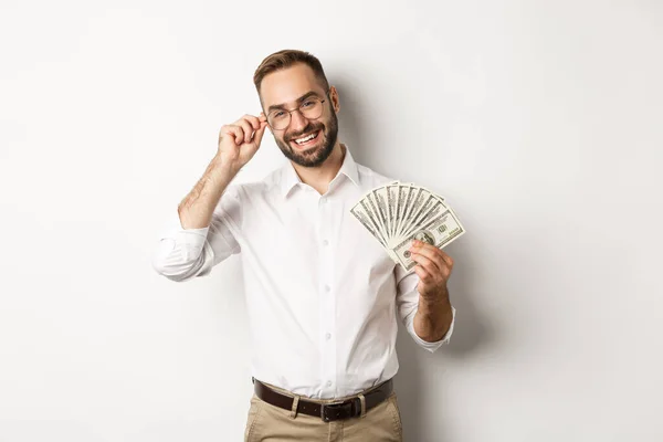 Bonito empresário bem sucedido segurando dinheiro, fixando óculos no nariz, de pé sobre fundo branco — Fotografia de Stock