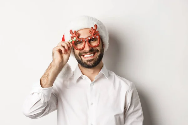 Крупный план красивого бородатого парня в рождественских очках и шляпе Санты, улыбающегося и желающего счастливого Рождества, стоящего на белом фоне — стоковое фото