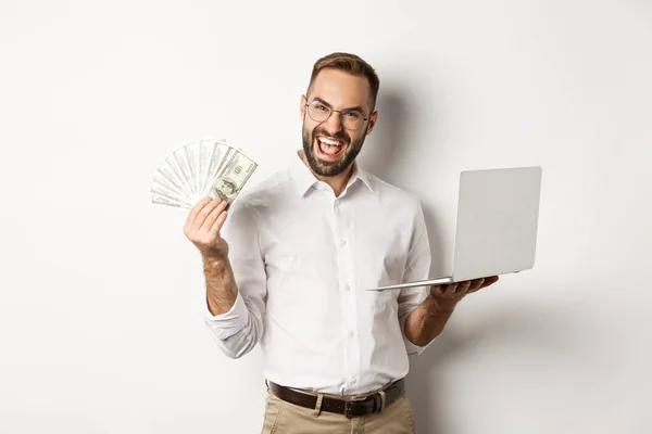 Negocios y comercio electrónico. Emocionado hombre de negocios sosteniendo dinero dólares y portátil, trabajando en línea, de pie sobre fondo blanco — Foto de Stock