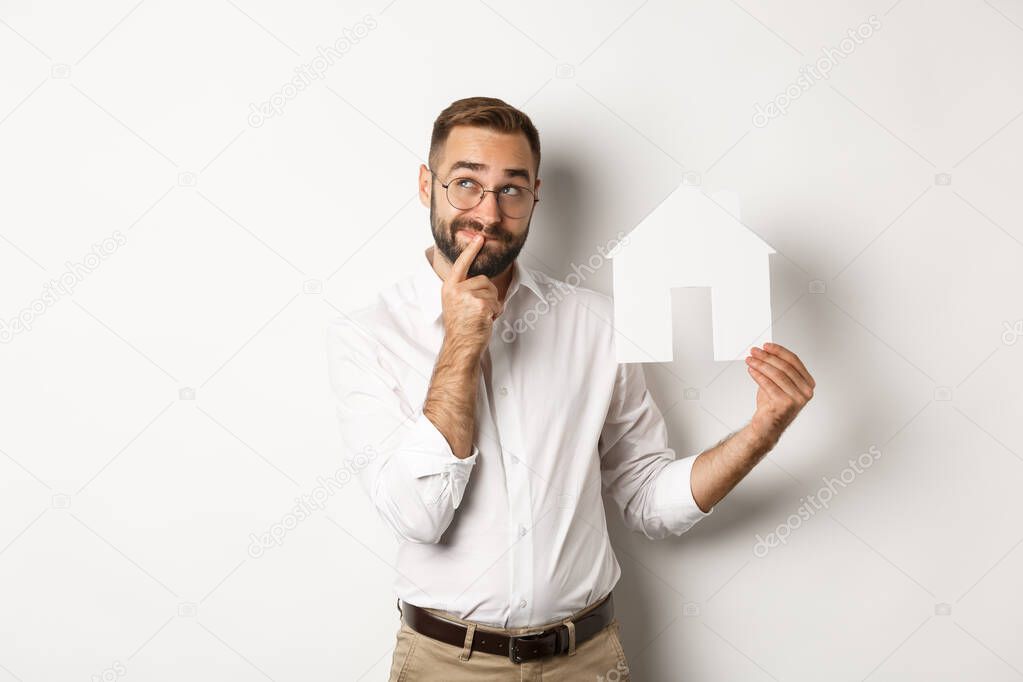 Real estate. Man thinking while searching for apartment, holding paper house model, standing over white background.