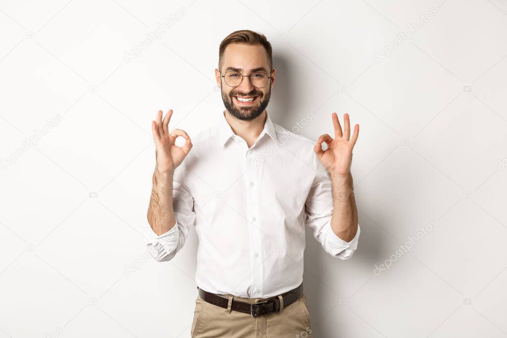 Satisfied handsome businessman showing ok sign, gurantee quallity, standing pleased against white background