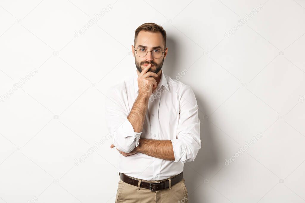 Thoughtful handsome businessman looking at camera, making choice or thinking, standing in glasses and white collar shirt against studio background
