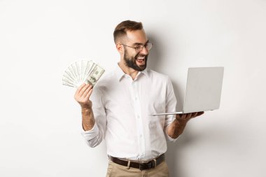 İş ve e-ticaret. Tatmin olmuş iş adamı dizüstü bilgisayarda iş yapıyor ve elinde para tutuyor, mutlu bir şekilde gülümsüyor, beyaz arka planda duruyor.