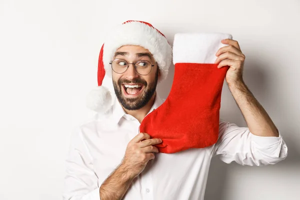 Close-up de homem feliz comemorando o Natal, receber presentes em meia xmas e olhando animado, vestindo chapéu de Papai Noel e óculos, fundo branco — Fotografia de Stock
