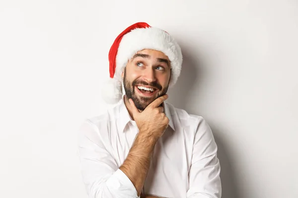 Fête, vacances d'hiver et concept de célébration. Gros plan de l'homme heureux qui planifie la liste des cadeaux de Noël, portant un chapeau de Père Noël, regardant le coin supérieur gauche réfléchi, fond blanc — Photo
