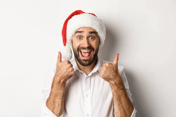 Party, Winterurlaub und Festkonzept. Mann genießt Weihnachten, trägt Weihnachtsmütze und zeigt Daumen hoch mit aufgeregtem Gesicht, weißer Hintergrund — Stockfoto