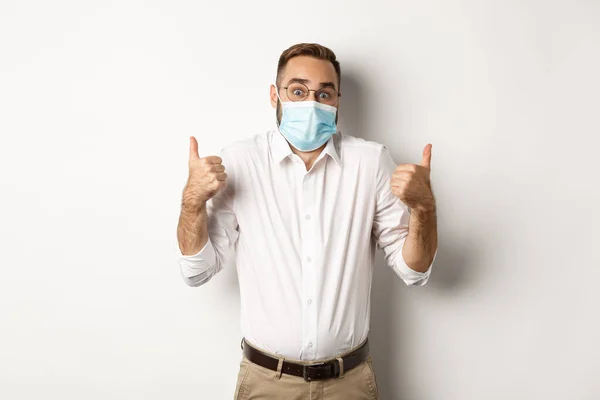 Covid-19, social distancing and quarantine concept. Impressed man in face mask showing thumbs-up and shrugging, standing amazed against white background — Stock Photo, Image