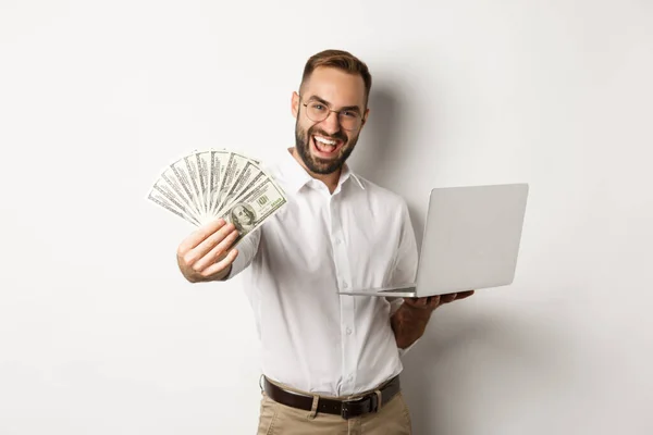 Negocios y comercio electrónico. Feliz empresario exitoso jactándose de dinero, trabajando en el ordenador portátil en línea, de pie sobre fondo blanco — Foto de Stock