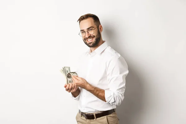 Homem de negócios bem sucedido contando dinheiro e sorrindo, de pé contra fundo branco e olhando satisfeito — Fotografia de Stock