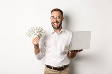 İş ve e-ticaret. Çevrimiçi, göz kırpan, para ve dizüstü bilgisayarın beyaz arka planda nasıl durduğunu gösteren kendine güvenen bir iş adamı.