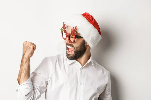 Close-up de homem barbudo feliz vestindo chapéu de Papai Noel e óculos de festa de Natal, fazendo punho bomba e alegria, alcançar objetivo e celebração, fundo branco — Fotografia de Stock