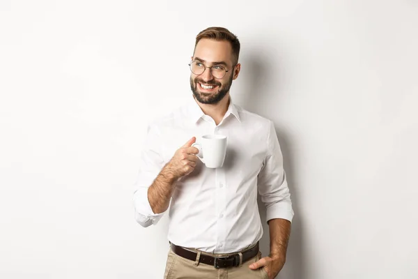 Framgångsrik affärsman dricker kaffe, tittar i sidled med nöjda leende, står över vit bakgrund — Stockfoto