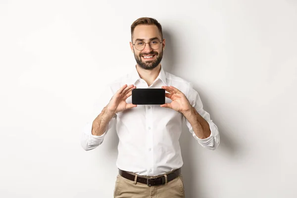 Snygg skäggig man i glasögon, demonstrera mobiltelefon ansökan, visar smartphone skärm, står över vit bakgrund — Stockfoto