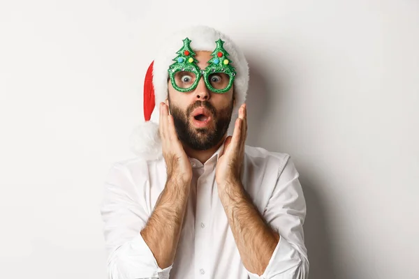 Homem adulto comemorando férias de inverno, vestindo óculos de festa de Natal e chapéu de Papai Noel, olhando surpreso para a câmera, em pé sobre fundo branco — Fotografia de Stock