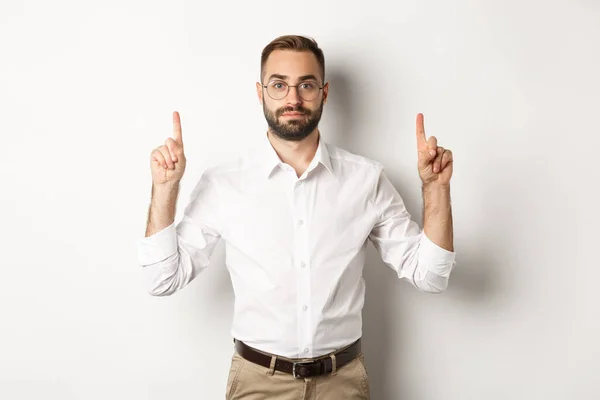 Jonge bebaarde zakenman wijzende vingers omhoog, tonen promo aanbod, staande over witte achtergrond — Stockfoto