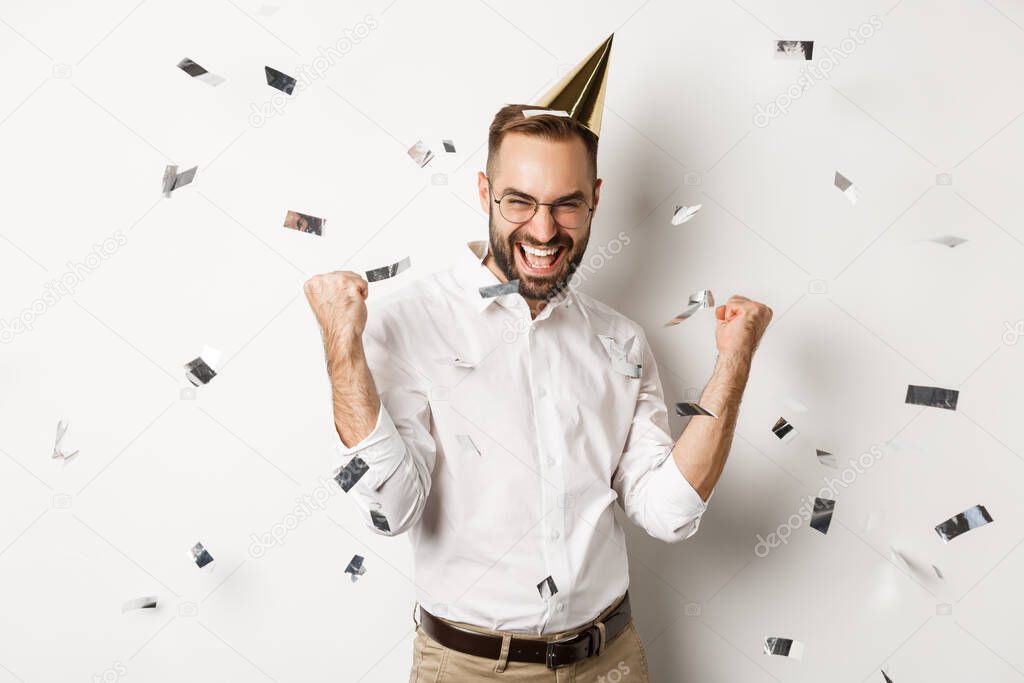 Celebration and holidays. Happy man dancing on birthday party with confetti, wearing b-day hat and rejoicing, standing over white background