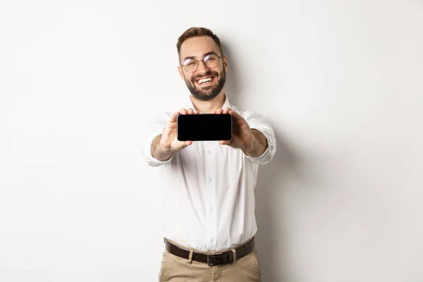 Glad affärsman visar mobilskärmen, håller telefonen horisontellt, står nöjd mot vit bakgrund — Stockfoto
