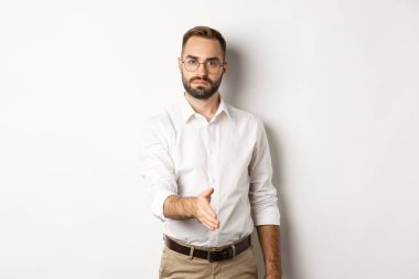 Kendine güvenen işadamı el sıkışmak için elini uzatıyor, iş ortağını selamlıyor, beyaz arka planda duruyor.