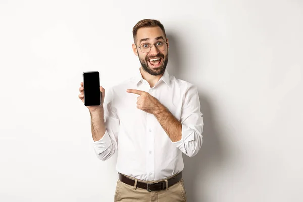 Beau mec excité montrant le téléphone portable, pointant du doigt l'écran et souriant, debout sur fond blanc — Photo