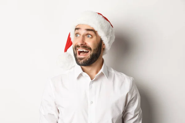 Primo piano di uomo barbuto stupito in cappello di Babbo Natale, guardando a sinistra con eccitazione, offerta promozionale di Natale, in piedi su sfondo bianco — Foto Stock