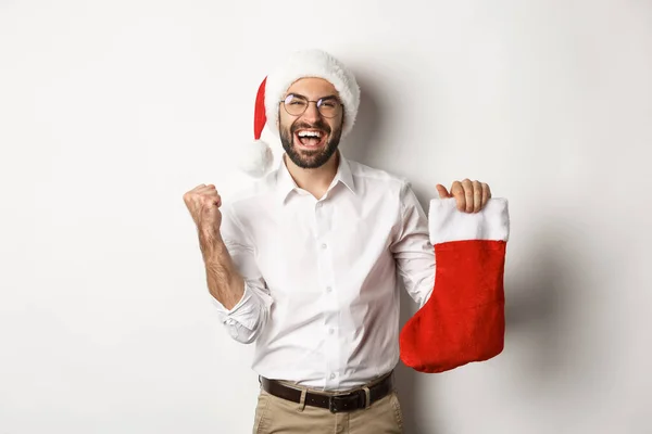 Frohe Weihnachten, Feiertage Konzept. Glücklicher erwachsener Mann erhält Geschenke in Weihnachtssocke, sieht aufgeregt aus, trägt Weihnachtsmütze, weißer Hintergrund — Stockfoto