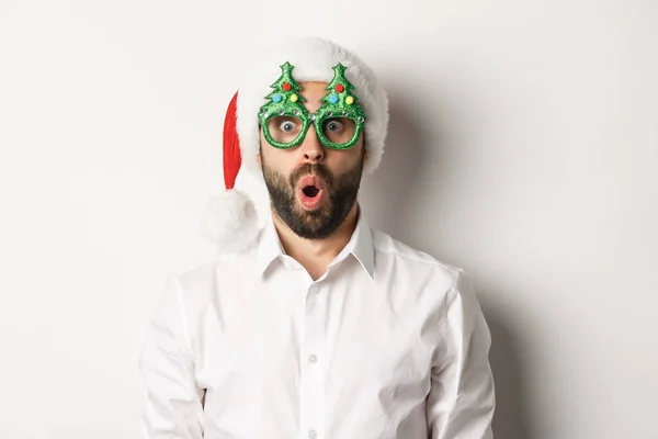 Homem adulto comemorando férias de inverno, vestindo óculos de festa de Natal e chapéu de Papai Noel, olhando surpreso para a câmera, em pé sobre fundo branco — Fotografia de Stock