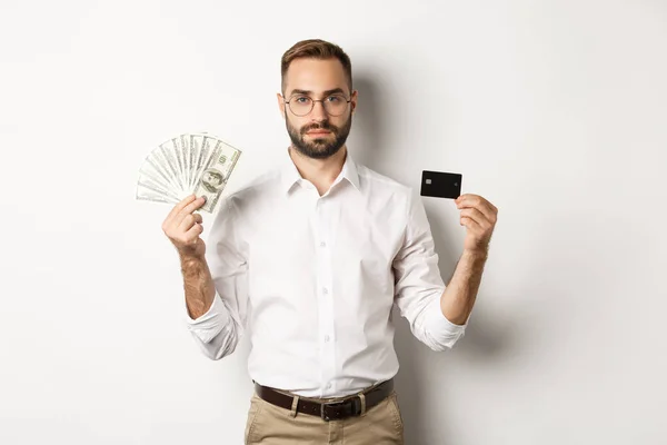 Homem de negócios sério olhando para a câmera, segurando cartão de crédito e dinheiro, em pé sobre fundo branco. Conceito de compras e finanças — Fotografia de Stock