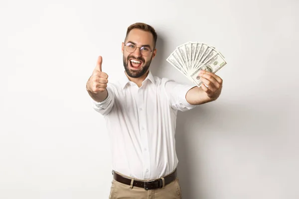 Homem animado mostrando polegares para cima e dinheiro, ganhando dinheiro, de pé sobre fundo branco — Fotografia de Stock
