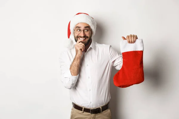 Веселого Різдва, концепція свят. Дорослий чоловік виглядає щасливим і допитливим на різдвяній шкарпетці, отримує подарунки, одягнені в капелюх Санти, білий фон — стокове фото