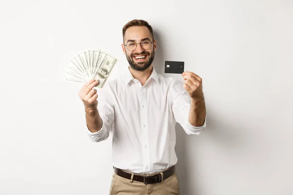 Jovem feliz mostrando seu cartão de crédito e dinheiro dólares, sorrindo satisfeito, de pé sobre fundo branco — Fotografia de Stock