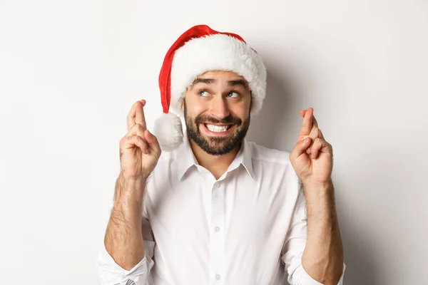 Party, Winterurlaub und Festkonzept. Glücklicher Mann mit Weihnachtsmütze, Weihnachtswunsch, Daumen drücken für viel Glück und aufgeregtem Lächeln, weißer Hintergrund — Stockfoto