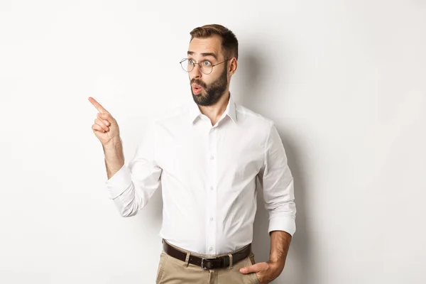 Geïntrigeerd jonge man met bril wijzend vinger links, op zoek naar promo aanbod geïnteresseerd, staande tegen een witte achtergrond — Stockfoto