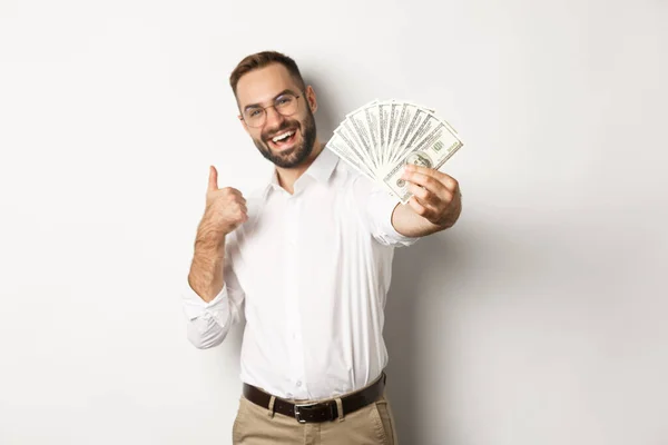 Exitoso hombre de negocios mostrando dinero dólares y pulgares hacia arriba, sonriendo satisfecho, de pie sobre fondo blanco — Foto de Stock