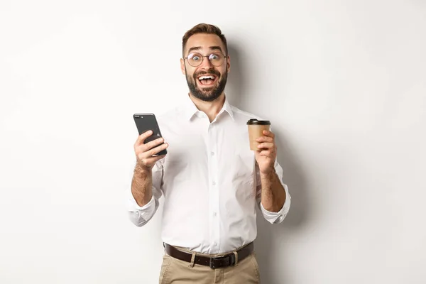 Homme d'affaires incroyable buvant du café, réagissant à une offre en ligne impressionnante sur téléphone mobile, debout sur fond blanc — Photo