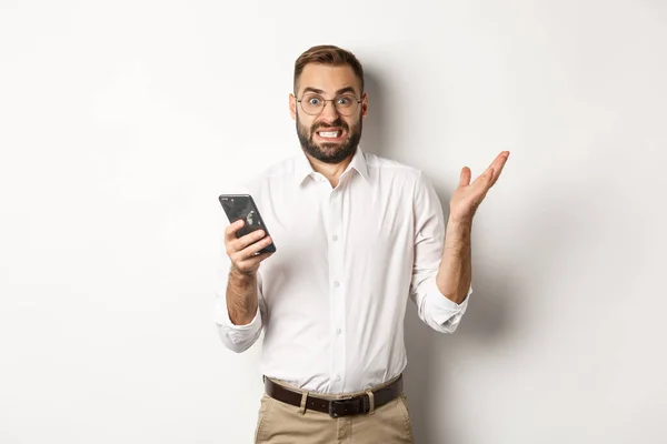 Homme d'affaires confus lisant un message étrange sur un téléphone portable, regardant agacé, debout sur fond blanc — Photo