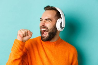 Kulaklıkla müzik dinleyen, turuncu süveter giymiş turkuaz arka planda duran yakışıklı modern adamın yakın çekimi.