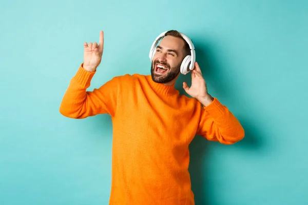 Turuncu süveter giyen mutlu yetişkin adam, yukarı bakıyor ve kulaklıkla müzik dinliyor, mavi arka planda duruyor. — Stok fotoğraf