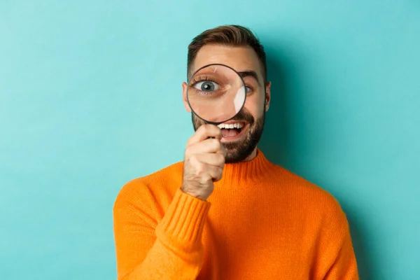 Uomo divertente guardando attraverso lente d'ingrandimento, ricerca o indagare qualcosa, in piedi in maglione arancione contro sfondo turchese — Foto Stock