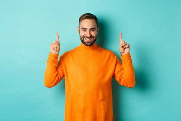 Beau mec barbu en pull orange apparaissant promo, pointant vers le haut et souriant, debout sur fond turquoise — Photo