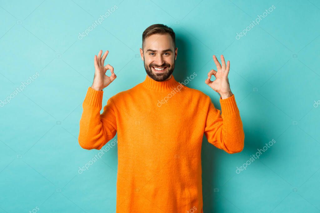 Image of confident smiling man showing okay sign, approve and agree, guarantee quality, standing over light blue background.