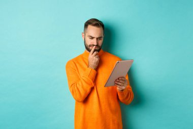 Ciddi bir adam dijital tablet ekranı inceliyor, sosyal medya okuyor, turkuaz arka planda dikiliyor.