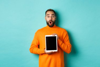 Teknoloji. Şaşırmış sakallı adam çenesini indirir, kameraya bakar ve dijital tablet ekranı gösterir, açık mavi arkaplan