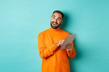 Yakışıklı bir adam düşünüyor ve dijital tablet kullanıyor, online alışveriş yapıyor ve fikirleri inceliyor, sol üst köşeye bakıyor, açık mavi arkaplanın üzerinde duruyor.