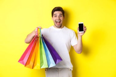 Alışveriş torbaları tutan ve cep telefonu ekranını gösteren genç adam, para başvurusu, sarı arka planda duruyor.