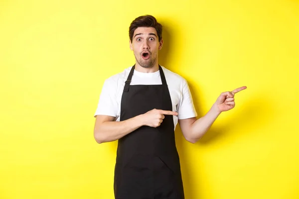 Дивовижний офіціант в чорному фартусі показує промо-пропозицію, вказуючи пальці праворуч і виглядаючи здивованим, стоїть в уніформі на жовтому тлі — стокове фото