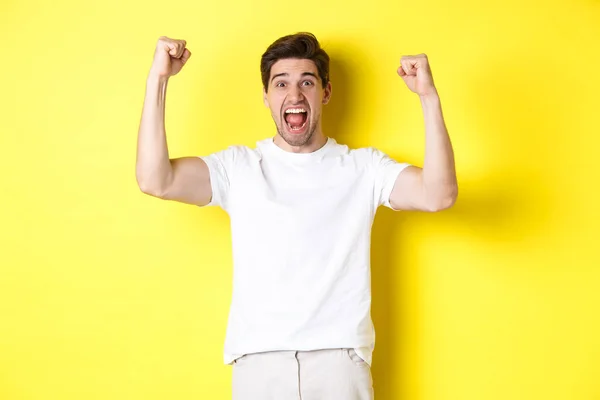 Imagen del hombre excitado ganando, levantando las manos y celebrando, triunfando y enraizándose por el equipo, de pie sobre el fondo amarillo — Foto de Stock