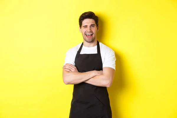 Bonito barista de avental preto piscando para você, vestindo uniforme de avental preto, de pé sobre fundo amarelo — Fotografia de Stock