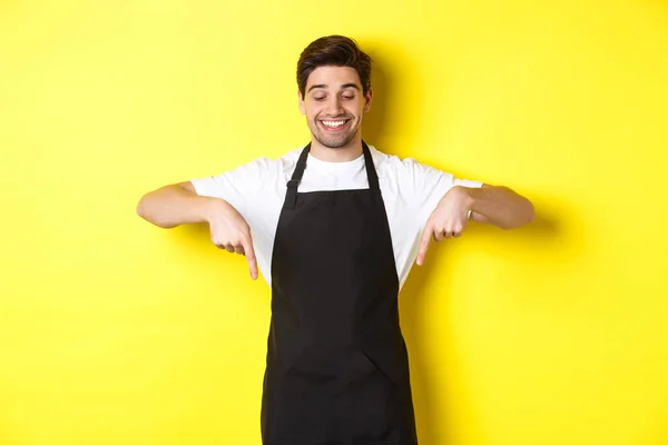 Glad café arbetare i svart förkläde pekar finger ner, ser glad och ler, visar logotyp, står över gul bakgrund — Stockfoto