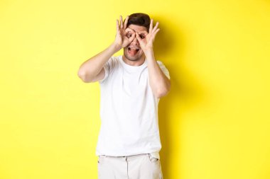 Genç adam komik yüzler ve yapışkan dil gösteriyor, sarı arka planda şakacı duruyor.